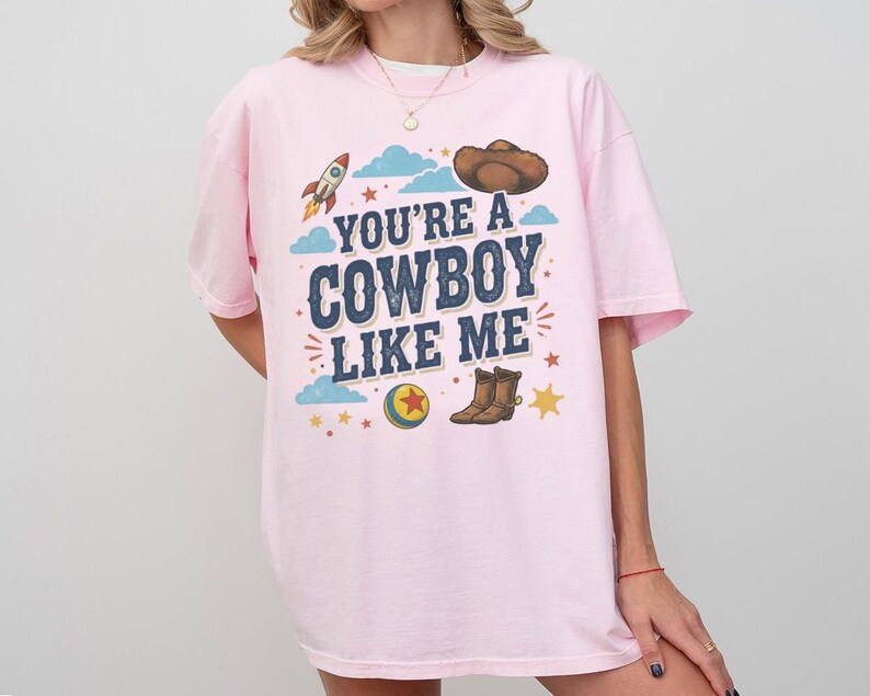 May include: A light pink t-shirt featuring the dark blue text "YOU'RE A COWBOY LIKE ME" printed on the chest. The graphic includes a brown cowboy hat, a rocket ship, a colourful ball with a red star, brown cowboy boots, and small yellow stars.