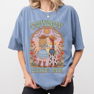 Puede incluir: Una camiseta azul polvoriento con un estampado gr&aacute;fico de dos figuras con sombreros de vaquero dentro de un arco floral. El texto COWBOY LIKE ME est&aacute; impreso en letras verdes sobre y debajo de la ilustraci&oacute;n, que incluye amapolas y margaritas.