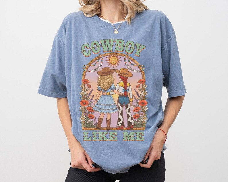 Puede incluir: Una camiseta azul polvoriento con un estampado gr&aacute;fico de dos figuras con sombreros de vaquero dentro de un arco floral. El texto COWBOY LIKE ME est&aacute; impreso en letras verdes sobre y debajo de la ilustraci&oacute;n, que incluye amapolas y margaritas.