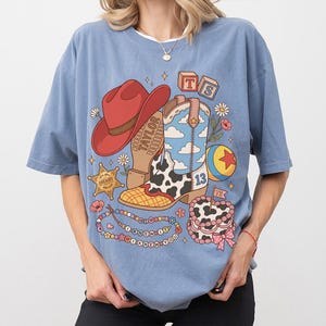 May include: A dusty blue t-shirt featuring a graphic print of a red cowboy hat, a cow-print boot with a blue sky and cloud pattern, a sheriff star, a colourful ball, a beaded necklace with text, and a cake with the text TS and the number 13.