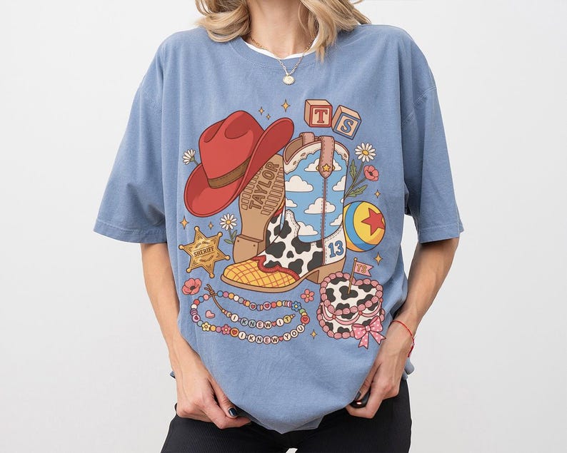 May include: A dusty blue t-shirt featuring a graphic print of a red cowboy hat, a cow-print boot with a blue sky and cloud pattern, a sheriff star, a colourful ball, a beaded necklace with text, and a cake with the text TS and the number 13.