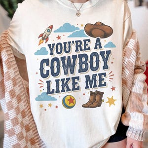 May include: A cream-coloured t-shirt featuring a graphic print with the text "YOU'RE A COWBOY LIKE ME" in dark blue letters. The design includes a brown cowboy hat, a rocket, a colourful ball with a red star, brown cowboy boots, and blue clouds.