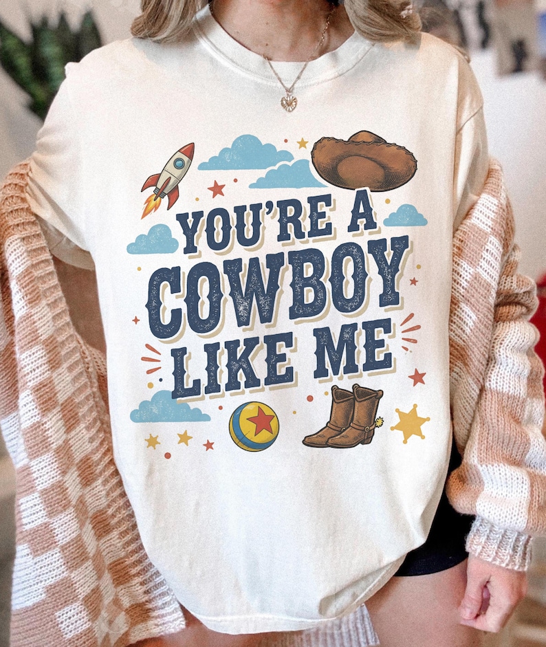 May include: A cream-coloured t-shirt featuring a graphic print with the text "YOU'RE A COWBOY LIKE ME" in dark blue letters. The design includes a brown cowboy hat, a rocket, a colourful ball with a red star, brown cowboy boots, and blue clouds.