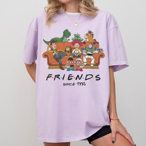 K&ouml;nnte beinhalten: Ein hellviolettes Kurzarm-T-Shirt mit einer Grafik verschiedener Zeichentrickfiguren auf einem orangefarbenen Sofa. Unter der Grafik steht der Text F.R.I.E.N.D.S mit bunten Punkten zwischen den Buchstaben und dem Text SINCE 1995 darunter.
