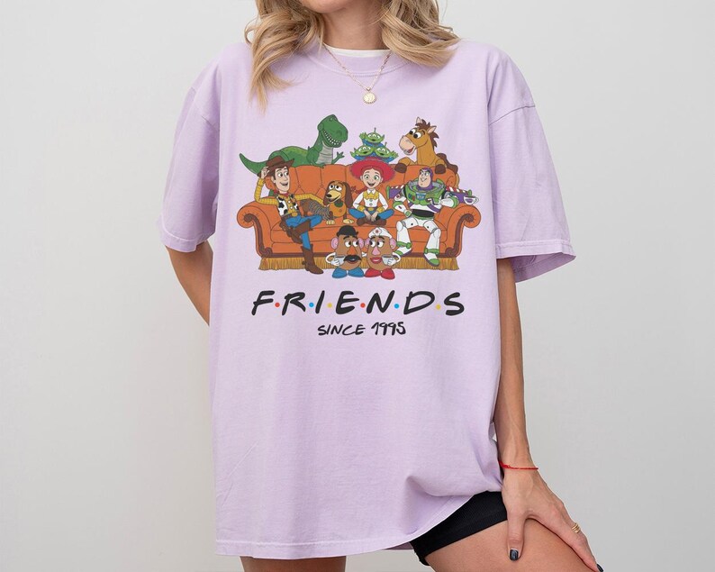 K&ouml;nnte beinhalten: Ein hellviolettes Kurzarm-T-Shirt mit einer Grafik verschiedener Zeichentrickfiguren auf einem orangefarbenen Sofa. Unter der Grafik steht der Text F.R.I.E.N.D.S mit bunten Punkten zwischen den Buchstaben und dem Text SINCE 1995 darunter.