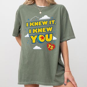 Pode incluir: Uma t-shirt verde-oliva com o texto amarelo I KNEW IT, I KNEW YOU, onde a letra O &eacute; substitu&iacute;da por um gr&aacute;fico de bola azul, amarela e vermelha. Uma etiqueta vermelha em forma de cora&ccedil;&atilde;o com as letras TS pende abaixo, rodeada de nuvens.