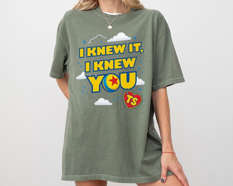 Pode incluir: Uma t-shirt verde-oliva com o texto amarelo I KNEW IT, I KNEW YOU, onde a letra O &eacute; substitu&iacute;da por um gr&aacute;fico de bola azul, amarela e vermelha. Uma etiqueta vermelha em forma de cora&ccedil;&atilde;o com as letras TS pende abaixo, rodeada de nuvens.