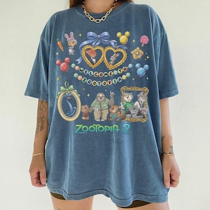 May include: Blue Zootopia 2 graphic t-shirt. The shirt features cartoon characters and the text "WELCOME TO ZOOTOPIA" and "ZOOTOPIA 2". The design includes hearts, Mickey Mouse heads, and other decorative elements. The shirt has short sleeves.