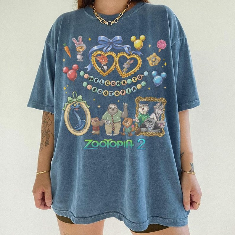 May include: Blue Zootopia 2 graphic t-shirt. The shirt features cartoon characters and the text "WELCOME TO ZOOTOPIA" and "ZOOTOPIA 2". The design includes hearts, Mickey Mouse heads, and other decorative elements. The shirt has short sleeves.