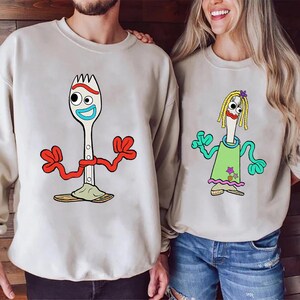 May include: Two beige crewneck sweatshirts featuring cartoon characters. The left sweatshirt displays a white spork character with red pipe cleaner arms and a blue mouth. The right sweatshirt shows a spork character with yellow yarn hair.