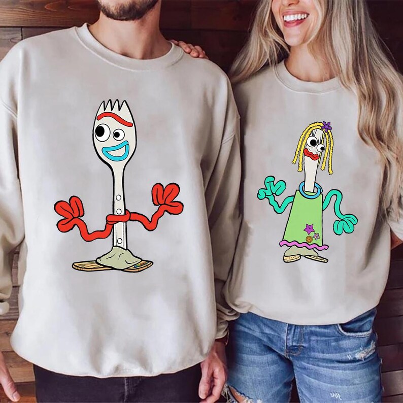 May include: Two beige crewneck sweatshirts featuring cartoon characters. The left sweatshirt displays a white spork character with red pipe cleaner arms and a blue mouth. The right sweatshirt shows a spork character with yellow yarn hair.
