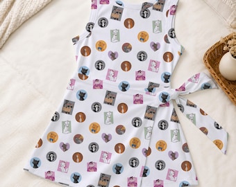 May include: A sleeveless white dress featuring a repeating pattern of various cartoon cats in colorful circles, squares, and hearts. The dress has a wrap-style front with a fabric tie at the waist, a round neckline, and a flared skirt silhouette.