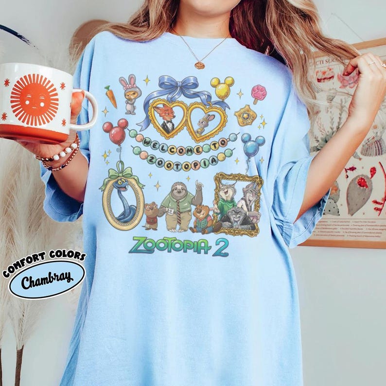 May include: Light blue t-shirt featuring Zootopia 2 graphics. The design includes framed character illustrations, Mickey Mouse-shaped balloons, and the text "WELCOME TO ZOOTOPIA". The shirt has a relaxed fit and is made of a soft material.