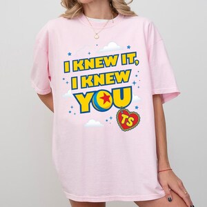 Pode incluir: Uma t-shirt rosa claro com o texto amarelo e azul I KNEW IT, I KNEW YOU, onde uma estrela vermelha numa bola azul e amarela substitui a letra O. Um pendente vermelho em forma de cora&ccedil;&atilde;o com as letras TS pende abaixo do texto.