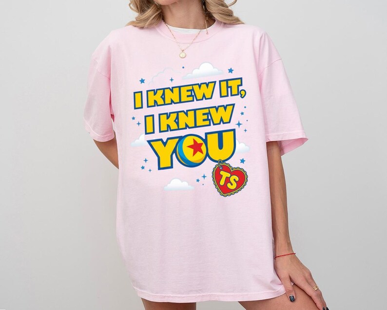 Pode incluir: Uma t-shirt rosa claro com o texto amarelo e azul I KNEW IT, I KNEW YOU, onde uma estrela vermelha numa bola azul e amarela substitui a letra O. Um pendente vermelho em forma de cora&ccedil;&atilde;o com as letras TS pende abaixo do texto.