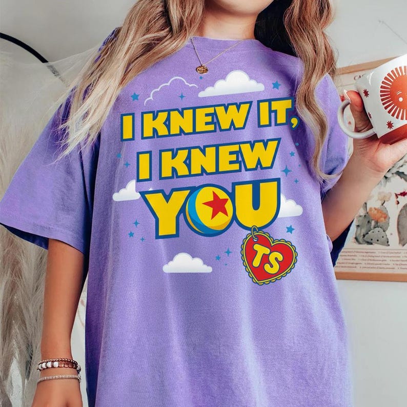 Pode incluir: Uma t-shirt roxa de manga curta com o texto amarelo I KNEW IT, I KNEW YOU, um gr&aacute;fico de uma bola com uma estrela vermelha e um pingente vermelho em forma de cora&ccedil;&atilde;o com as letras TS, rodeado por pequenas estrelas azuis e nuvens brancas.