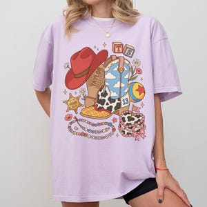 May include: A light purple t-shirt featuring a graphic print of a red cowboy hat, a blue and white cloud-patterned cowboy boot, a sheriff star, a colourful ball, a beaded necklace with text, and a small cake with the number 13 and a flag.