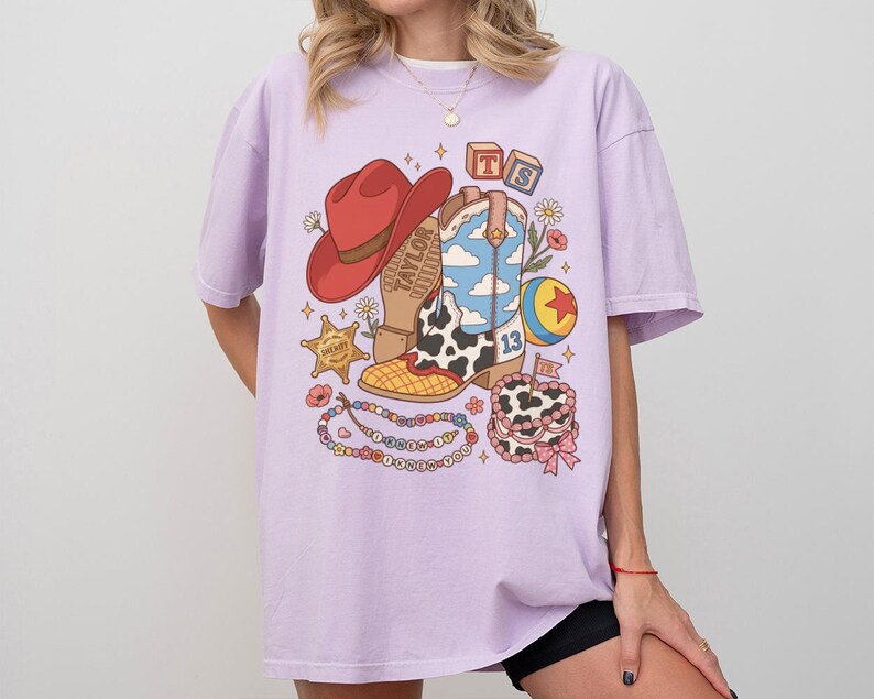 May include: A light purple t-shirt featuring a graphic print of a red cowboy hat, a blue and white cloud-patterned cowboy boot, a sheriff star, a colourful ball, a beaded necklace with text, and a small cake with the number 13 and a flag.