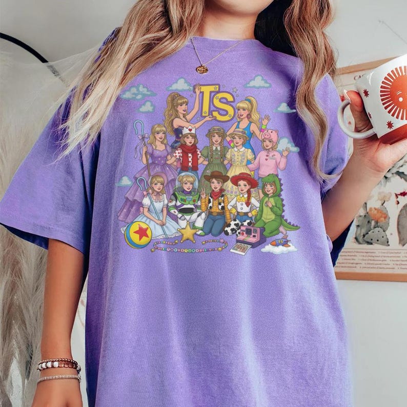 May include: A light purple short-sleeve t-shirt featuring a graphic print of various characters dressed in costumes inspired by the Toy Story franchise. The design includes the text TS at the top, a colorful ball, a star, and a camera illustration.