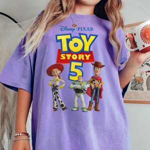 Op de afbeelding: Een lichtpaars t-shirt met het Disney Pixar Toy Story 5 logo in gele en rode tekst. De afbeelding toont de personages Jessie, Buzz Lightyear en Woody die zij aan zij in hun kenmerkende outfits op de voorkant van het shirt staan afgebeeld.