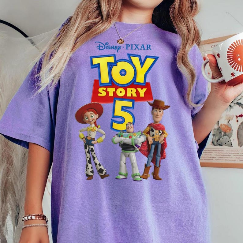 Op de afbeelding: Een lichtpaars t-shirt met het Disney Pixar Toy Story 5 logo in gele en rode tekst. De afbeelding toont de personages Jessie, Buzz Lightyear en Woody die zij aan zij in hun kenmerkende outfits op de voorkant van het shirt staan afgebeeld.