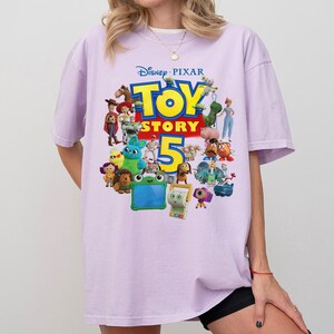 May include: A light purple short-sleeve t-shirt featuring a colorful graphic print of various Disney Pixar Toy Story characters and the text Toy Story 5. The design includes figures like Woody, Buzz Lightyear, and other animated characters.