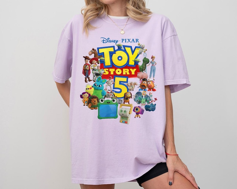 May include: A light purple short-sleeve t-shirt featuring a colorful graphic print of various Disney Pixar Toy Story characters and the text Toy Story 5. The design includes figures like Woody, Buzz Lightyear, and other animated characters.