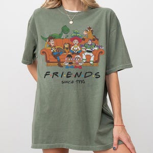 K&ouml;nnte beinhalten: Ein olivgr&uuml;nes Kurzarm-T-Shirt mit einem Grafikdruck verschiedener Zeichentrickfiguren, die auf einem orangefarbenen Sofa sitzen. Unter dem Sofa steht der Text FRIENDS in einer stilisierten Schrift mit bunten Punkten, gefolgt von SINCE 1995.