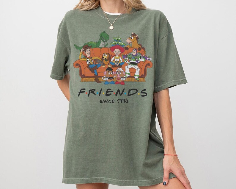 K&ouml;nnte beinhalten: Ein olivgr&uuml;nes Kurzarm-T-Shirt mit einem Grafikdruck verschiedener Zeichentrickfiguren, die auf einem orangefarbenen Sofa sitzen. Unter dem Sofa steht der Text FRIENDS in einer stilisierten Schrift mit bunten Punkten, gefolgt von SINCE 1995.