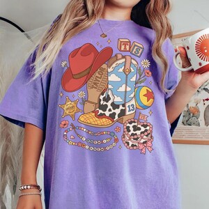 May include: A purple t-shirt featuring a graphic print of a red cowboy hat, a blue and white cow-print cowboy boot, a sheriff star, a colourful ball, a beaded necklace with the text I KNEW IT, and a cake with the text TS on a small flag.