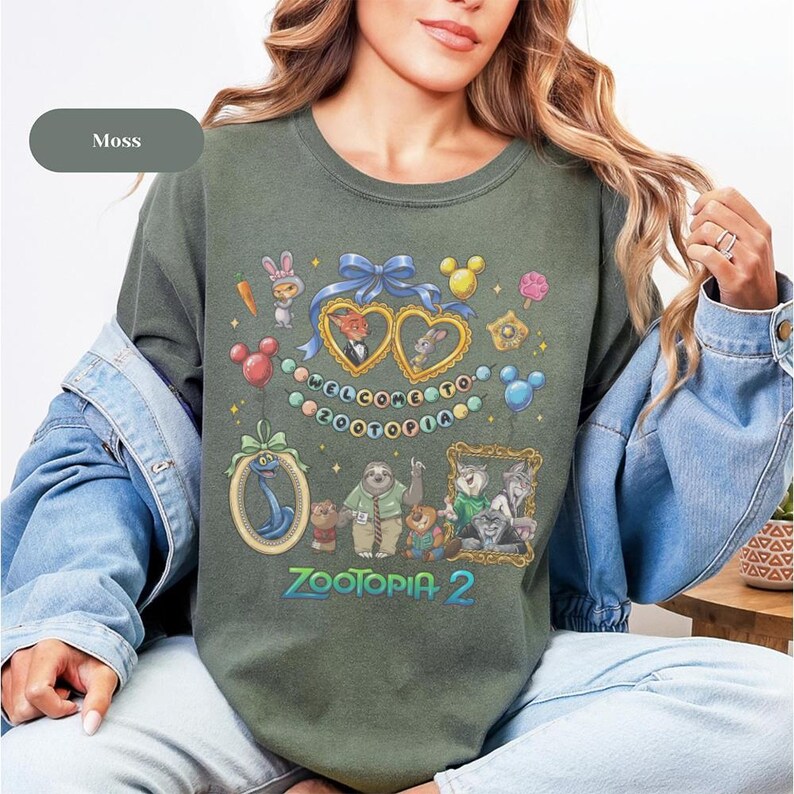 May include: Moss green crew neck sweatshirt featuring a colorful graphic from the movie Zootopia. The design includes characters, hearts, and the text "Welcome to Zootopia" and "Zootopia 2".