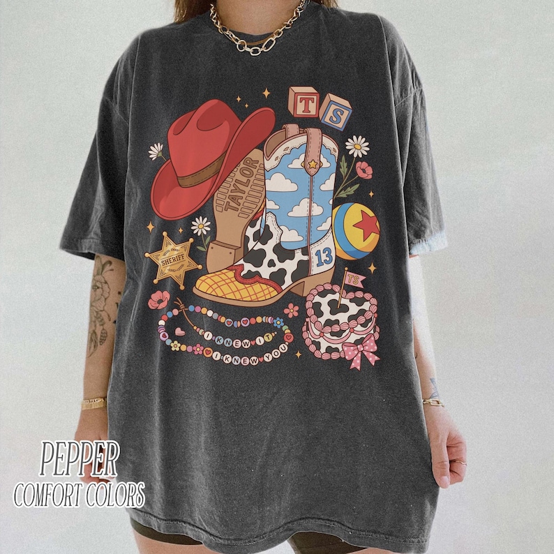 May include: A charcoal grey t-shirt featuring a graphic print of a red cowboy hat, a blue and white cloud-patterned cowboy boot with a cow print section, a yellow and red ball, a sheriff star, alphabet blocks, and a beaded necklace with text.