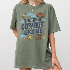 May include: An olive green short-sleeve t-shirt featuring a graphic print with the text "YOU'RE A COWBOY LIKE ME" in blue. The design includes a brown cowboy hat, a rocket, a colourful ball, brown boots, blue clouds, and yellow stars.
