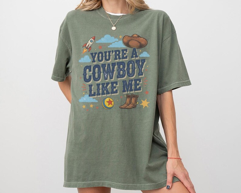 May include: An olive green short-sleeve t-shirt featuring a graphic print with the text "YOU'RE A COWBOY LIKE ME" in blue. The design includes a brown cowboy hat, a rocket, a colourful ball, brown boots, blue clouds, and yellow stars.