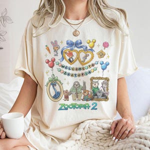 May include: Cream-colored t-shirt featuring a colorful graphic design with characters and text from the movie "Zootopia 2". The design includes framed portraits, balloons, and the words "WELCOME TO ZOOTOPIA".