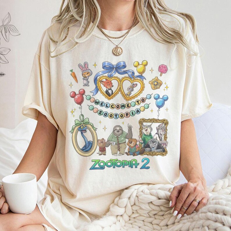 May include: Cream-colored t-shirt featuring a colorful graphic design with characters and text from the movie "Zootopia 2". The design includes framed portraits, balloons, and the words "WELCOME TO ZOOTOPIA".