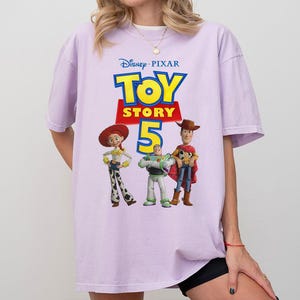 Op de afbeelding: Een lichtpaars T-shirt met korte mouwen en een grafische print van het Disney Pixar Toy Story 5-logo met de personages Jessie, Buzz Lightyear en Woody onder de tekst. Het shirt heeft een ronde hals en een ruimvallend ontwerp.