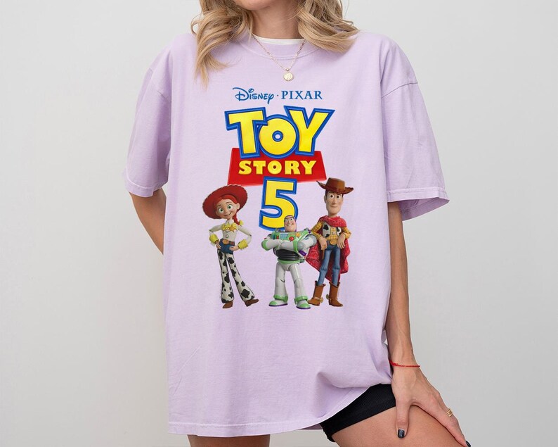 Op de afbeelding: Een lichtpaars T-shirt met korte mouwen en een grafische print van het Disney Pixar Toy Story 5-logo met de personages Jessie, Buzz Lightyear en Woody onder de tekst. Het shirt heeft een ronde hals en een ruimvallend ontwerp.