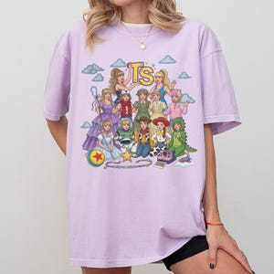 May include: A light purple short-sleeve t-shirt featuring a colorful graphic print of various animated characters and the letters TS at the top. The design includes multiple figures in diverse outfits, a yellow star, a ball, and a camera on a light background.
