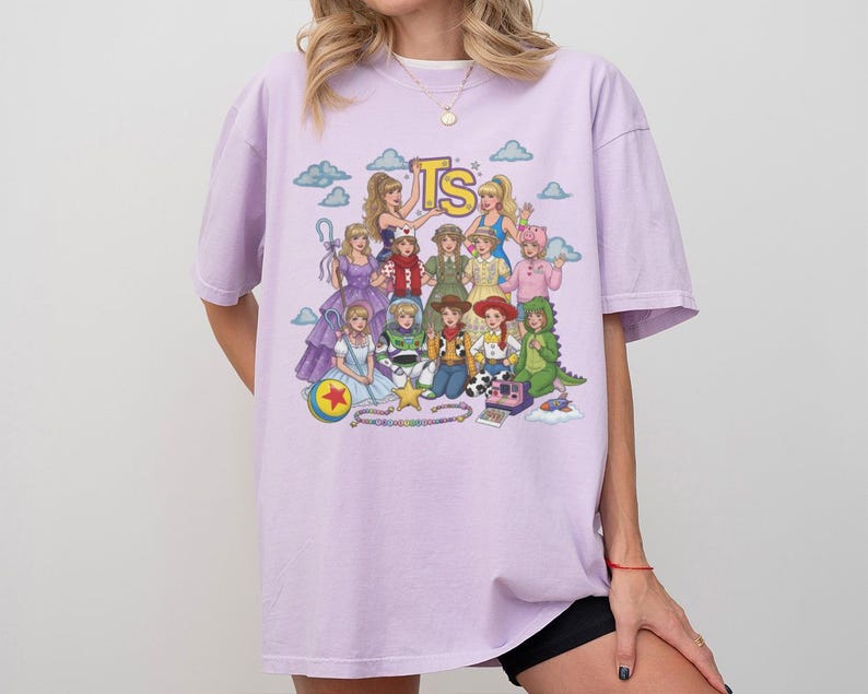 May include: A light purple short-sleeve t-shirt featuring a colorful graphic print of various animated characters and the letters TS at the top. The design includes multiple figures in diverse outfits, a yellow star, a ball, and a camera on a light background.
