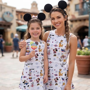 May include: Two sleeveless white dresses featuring a repeating pattern of cartoon characters, anchors, and cruise ships. Both individuals wear black mouse ear headbands. The child holds a colorful, round, multi-flavored ice cream treat on a stick.