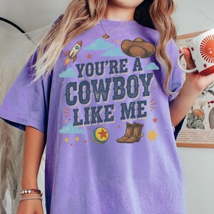 May include: A purple short-sleeve t-shirt featuring the text YOU'RE A COWBOY LIKE ME in dark blue letters. The graphic includes a rocket, a brown cowboy hat, a colourful ball with a red star, and brown cowboy boots surrounded by small yellow stars.