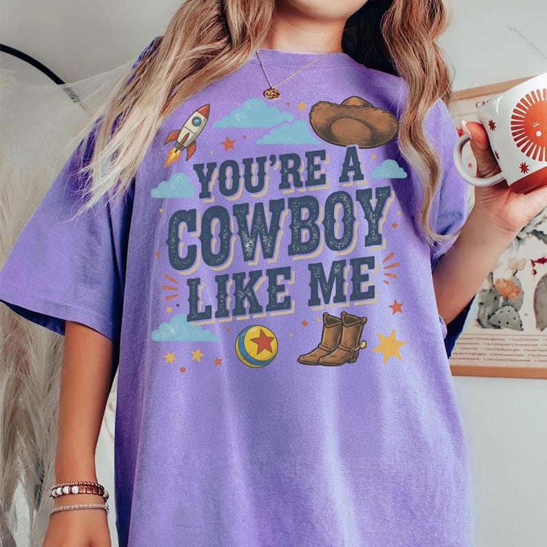 May include: A purple short-sleeve t-shirt featuring the text YOU'RE A COWBOY LIKE ME in dark blue letters. The graphic includes a rocket, a brown cowboy hat, a colourful ball with a red star, and brown cowboy boots surrounded by small yellow stars.
