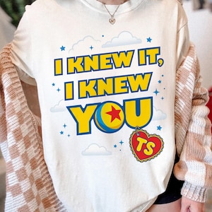 Pode incluir: Uma t-shirt cor de creme com o texto "I KNEW IT, I KNEW YOU" em letras amarelas em negrito com contornos azuis. O design inclui uma bola amarela e azul com uma estrela vermelha, um pingente em forma de cora&ccedil;&atilde;o com "TS" e estrelas e nuvens azuis.