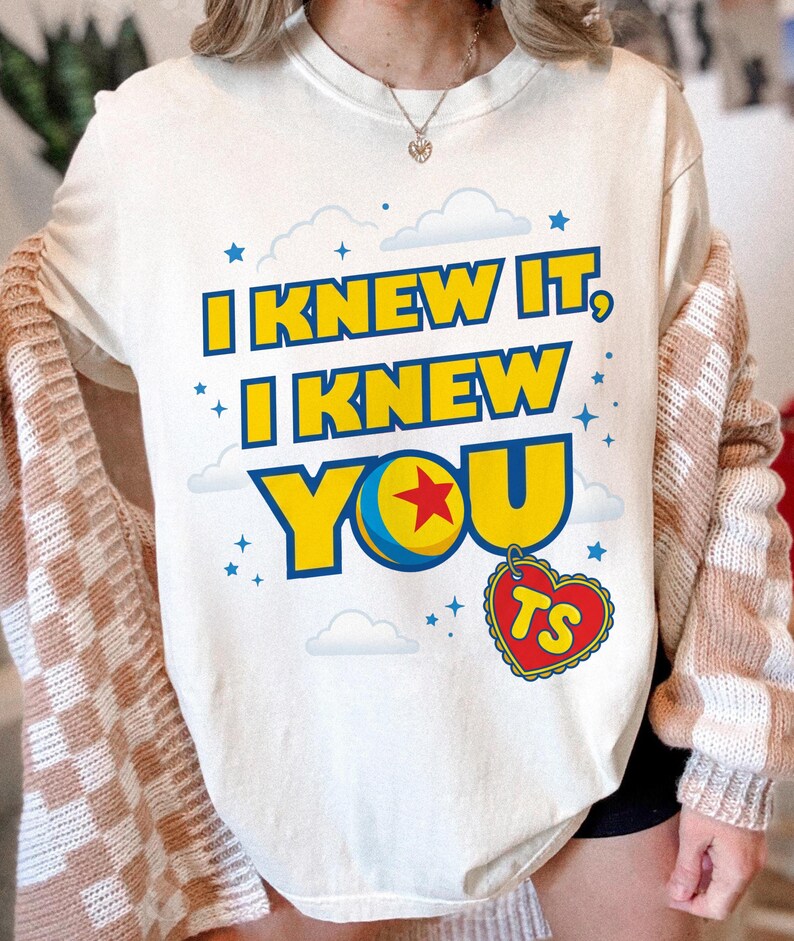 Pode incluir: Uma t-shirt cor de creme com o texto "I KNEW IT, I KNEW YOU" em letras amarelas em negrito com contornos azuis. O design inclui uma bola amarela e azul com uma estrela vermelha, um pingente em forma de cora&ccedil;&atilde;o com "TS" e estrelas e nuvens azuis.