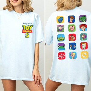 May include: A light blue t-shirt featuring the Disney Pixar Toy Story 5 logo on the front chest. The back displays a grid of fifteen colorful square icons depicting various characters and symbols from the Toy Story film franchise.