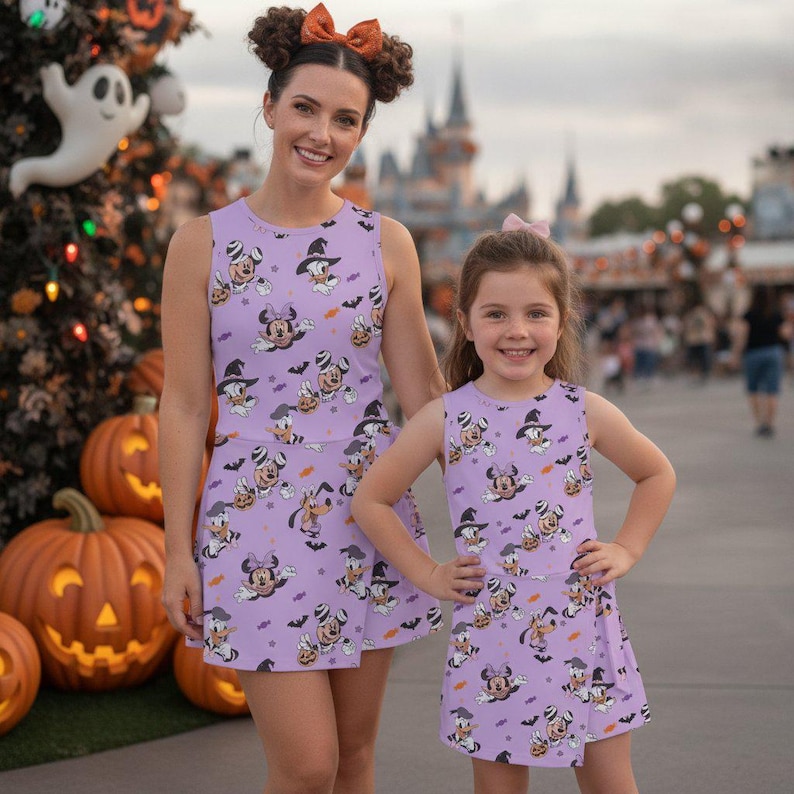 Może przedstawiać: Dwie sukienki bez rękaw&oacute;w w jasnofioletowym kolorze z nadrukiem postaci Disneya, w tym Myszki Minnie, Kaczki Daisy i Goofy'ego w kostiumach na Halloween, wraz z małymi dyniami, nietoperzami i ikonami cukierk&oacute;w w kształcie kukurydzy.