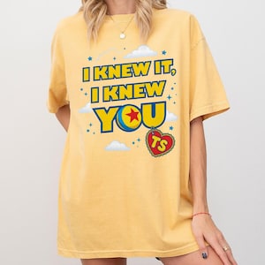 Pode incluir: Uma t-shirt de manga curta amarelo-p&aacute;lido com o texto azul e amarelo I KNEW IT, I KNEW YOU, um gr&aacute;fico de uma bola colorida com uma estrela vermelha, uma etiqueta vermelha em forma de cora&ccedil;&atilde;o com as letras TS e nuvens e estrelas brancas.