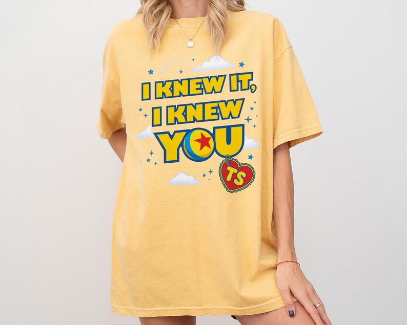 Pode incluir: Uma t-shirt de manga curta amarelo-p&aacute;lido com o texto azul e amarelo I KNEW IT, I KNEW YOU, um gr&aacute;fico de uma bola colorida com uma estrela vermelha, uma etiqueta vermelha em forma de cora&ccedil;&atilde;o com as letras TS e nuvens e estrelas brancas.