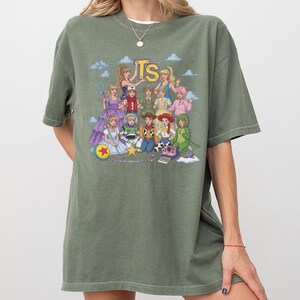 May include: An olive green short-sleeve t-shirt featuring a graphic print of various characters dressed in outfits inspired by Toy Story. The design includes the yellow text TS at the top, with characters arranged in a group against a blue sky background.